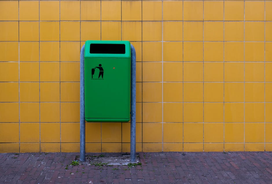 A green outdoor waste bin with a black icon depicting a person disposing of trash is mounted on two metal posts in front of a yellow tiled wall. The bin is positioned on a paved brick surface, with a