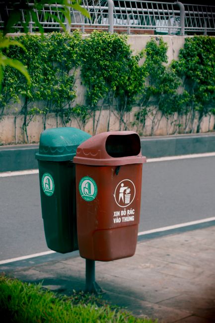 A green outdoor waste bin with a black icon depicting a person disposing of trash is mounted on two metal posts in front of a yellow tiled wall. The bin is positioned on a paved brick surface, with a