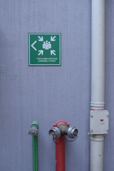 A green illuminated emergency exit sign featuring a running figure and an arrow pointing to the right is mounted on a dark industrial ceiling with exposed metal beams and ventilation pipes. The sign i